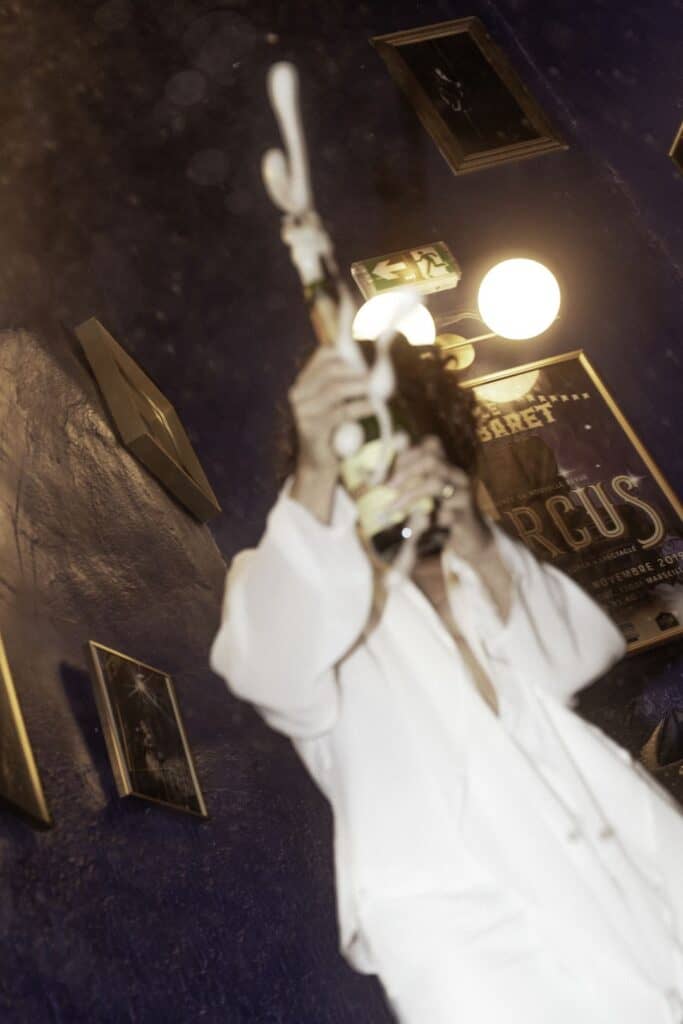Photo d'une artiste de cabaret en costume blanc, tenant un micro et une plume, dans une ambiance lum.